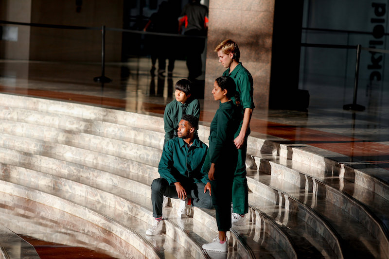 Four dancers, in evergreen jumpsuits, sit on the marble steps of Brookfield Place.